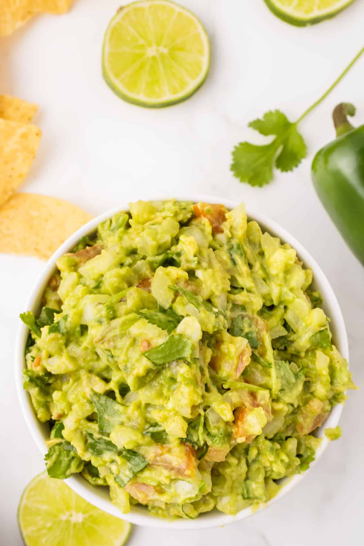 Easy guacamole in a white serving bowl on a marble countertop accented by tortilla chips, limes and jalapeños.