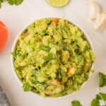 Easy guacamole in a white serving bowl on a marble countertop with assorted guacamole ingredients.