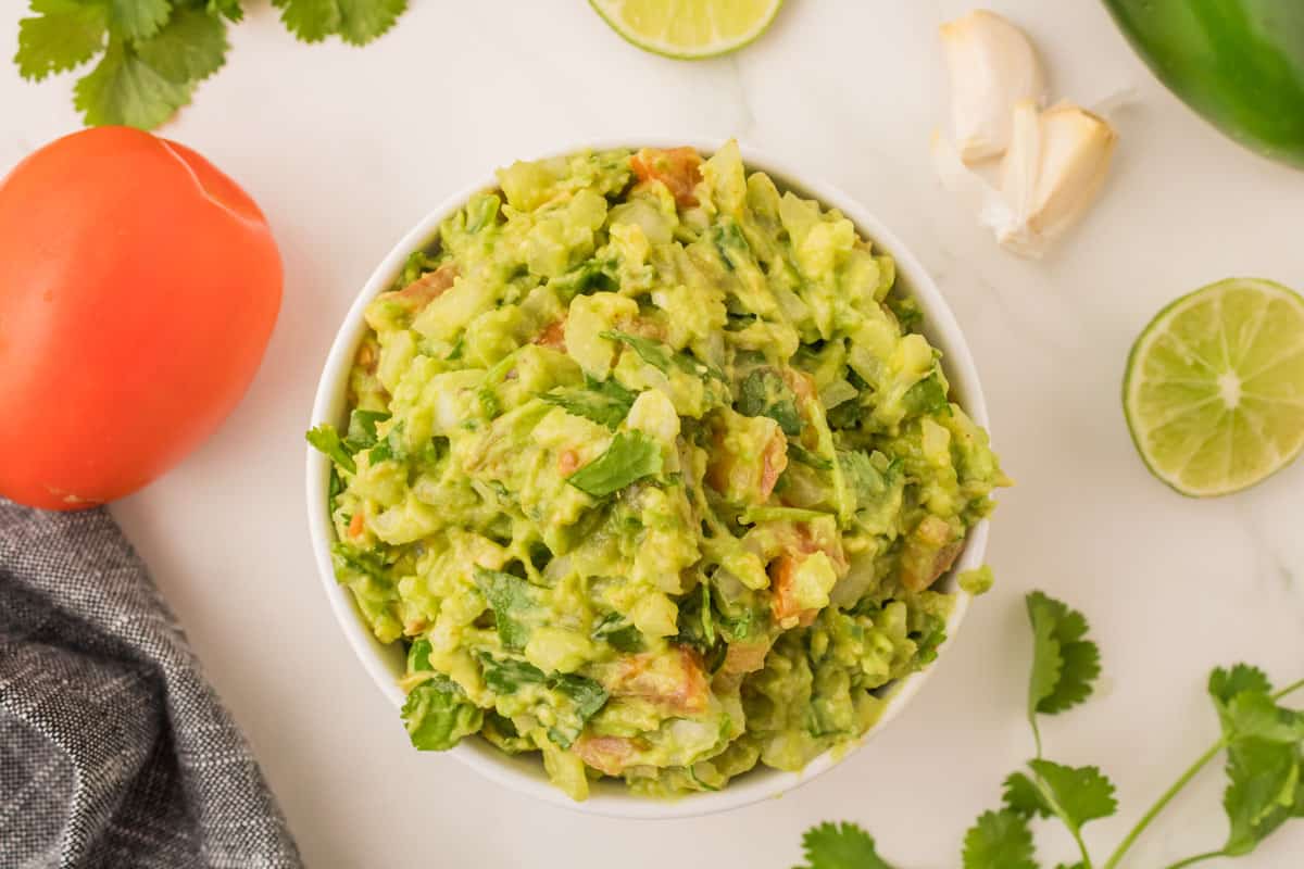 Easy guacamole in a white serving bowl on a marble countertop with assorted guacamole ingredients.