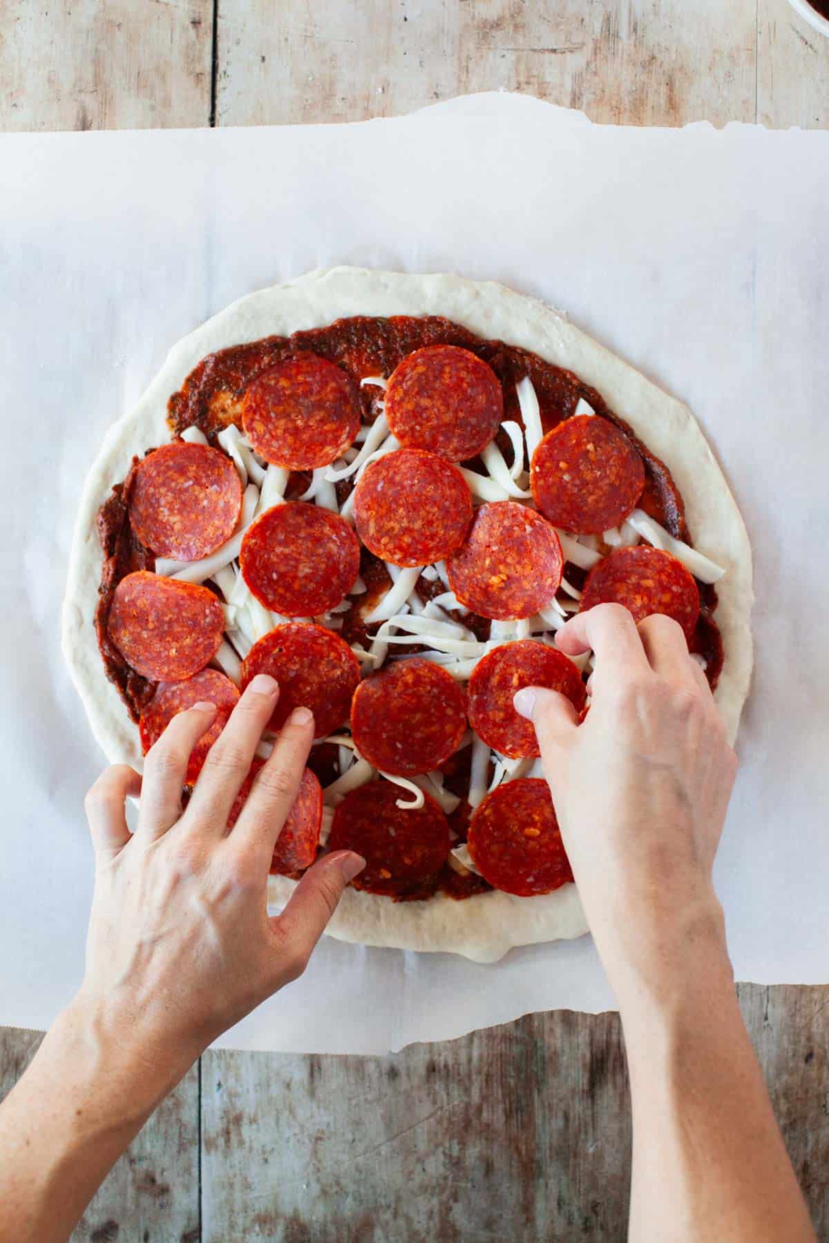 A set of hands placing sliced pepperoni on top of pizza dough with pizza sauce and shredded mozzarella.