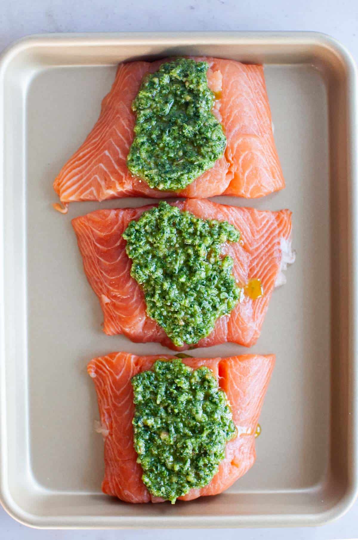 Three raw salmon filets topped with pesto on a unlined baking sheet.