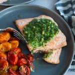 The corner of a pesto salmon filet cut off with a fork with roasted tomatoes on a gray plate.