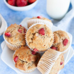 Overhead photo of multiple strawberry muffins arranged on a small white plate with a glass bottle filled with milk and a small bowl of whole strawberries.