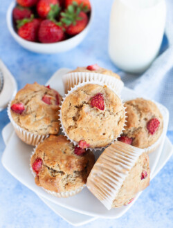 Overhead photo of multiple strawberry muffins arranged on a small white plate with a glass bottle filled with milk and a small bowl of whole strawberries.
