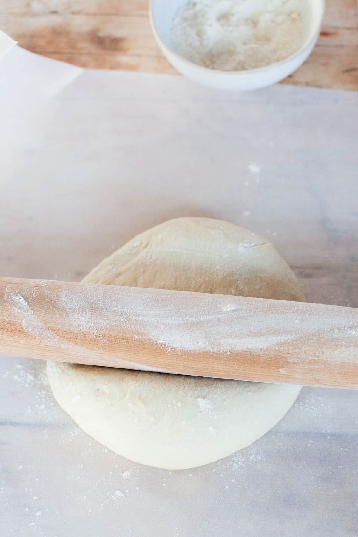 Thin crust pizza dough on parchment paper being rolled out with wooden rolling pin.