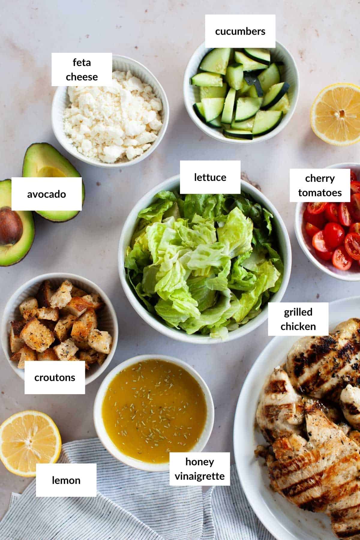 Bowls and plates of grilled chicken salad&mdash;featuring lettuce, cucumbers, cherry tomatoes, feta cheese, avocado, croutons, lemon, and honey vinaigrette&mdash;are arranged on a tabletop with labels identifying each ingredient.