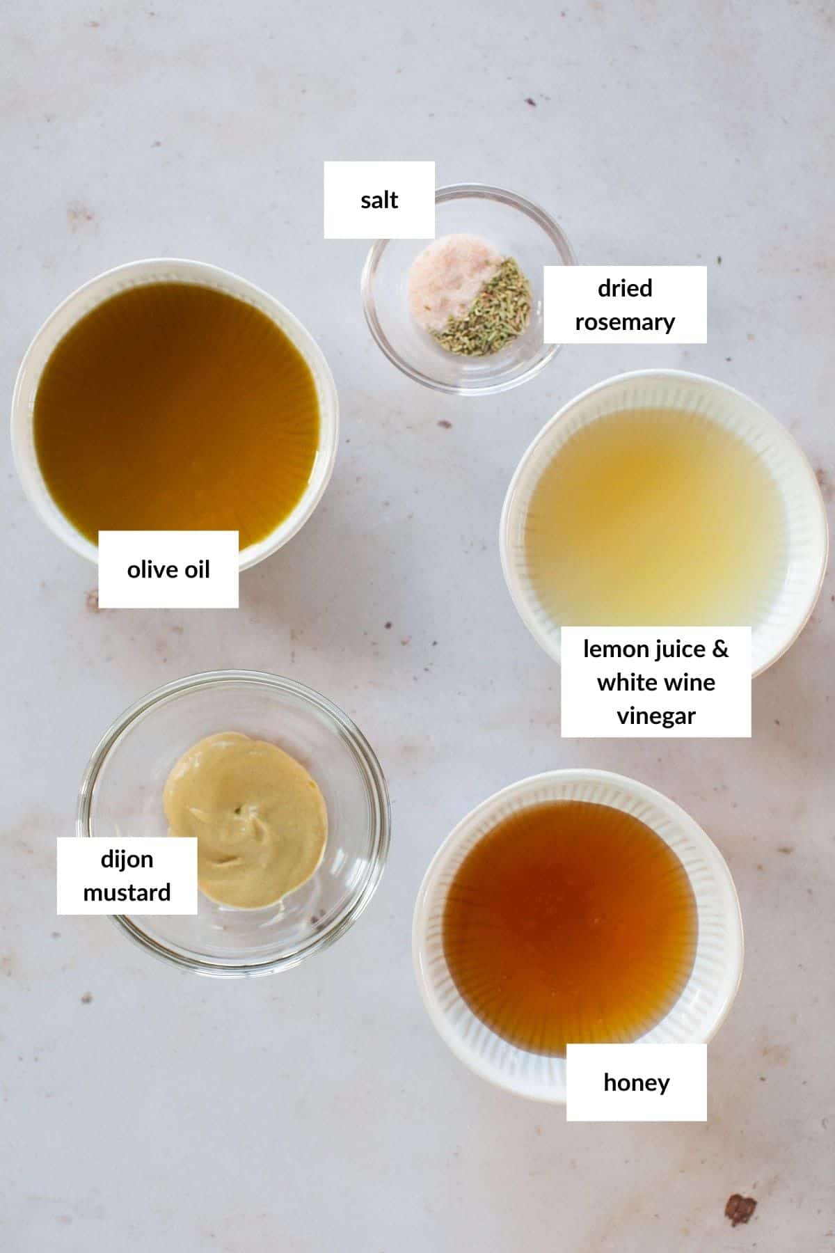 Five labeled bowls containing olive oil, salt, dried rosemary, lemon juice with white wine vinegar, Dijon mustard, and honey are arranged on a light surface&mdash;perfect ingredients for a flavorful grilled chicken salad.