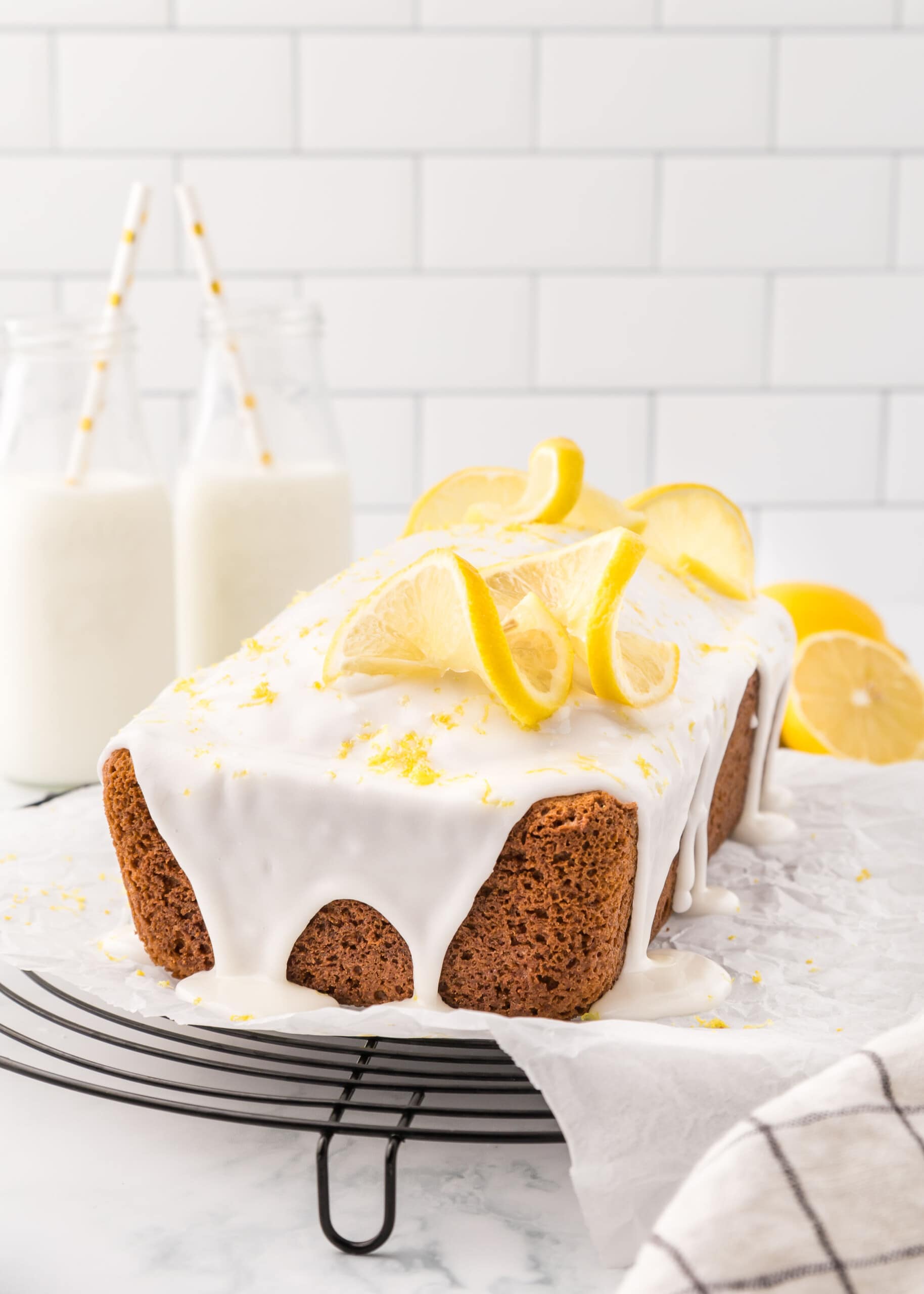 Lemon loaf with icing sprinkled with lemon zest and topped with two twisted lemon slices accented with glass milk bottles with white and yellow straws.