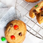 M&M cookie leaning against a wire cooling rack with an assortment of M&M's surrounding the cookie.