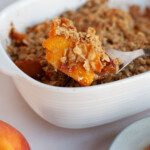 A small serving of peach crisp being spooned out of a white serving dish.