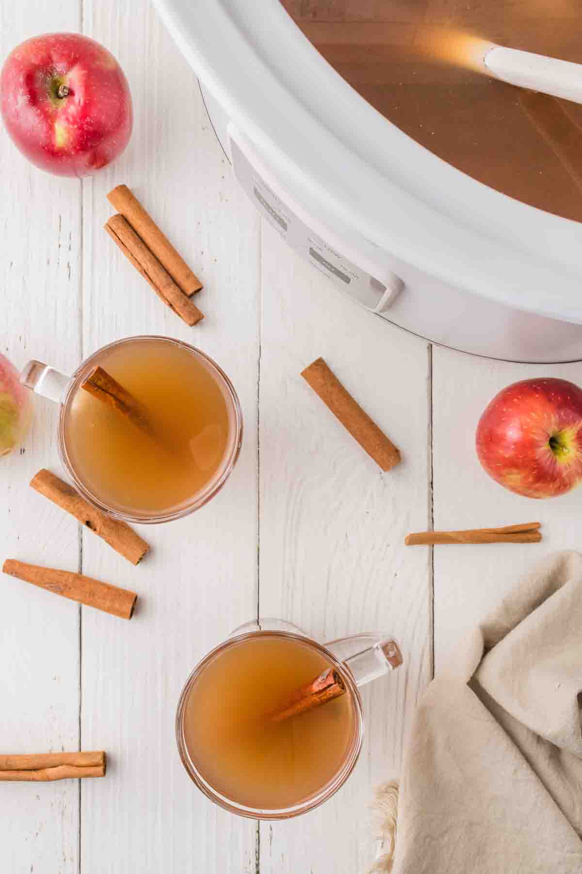 Spiced apple cider in two clear glasses with a cinnamon stick on a white, wooden table.