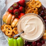 Fruit dip in a glass bowl surrounded by a platter of assorted fruits and pretzels.