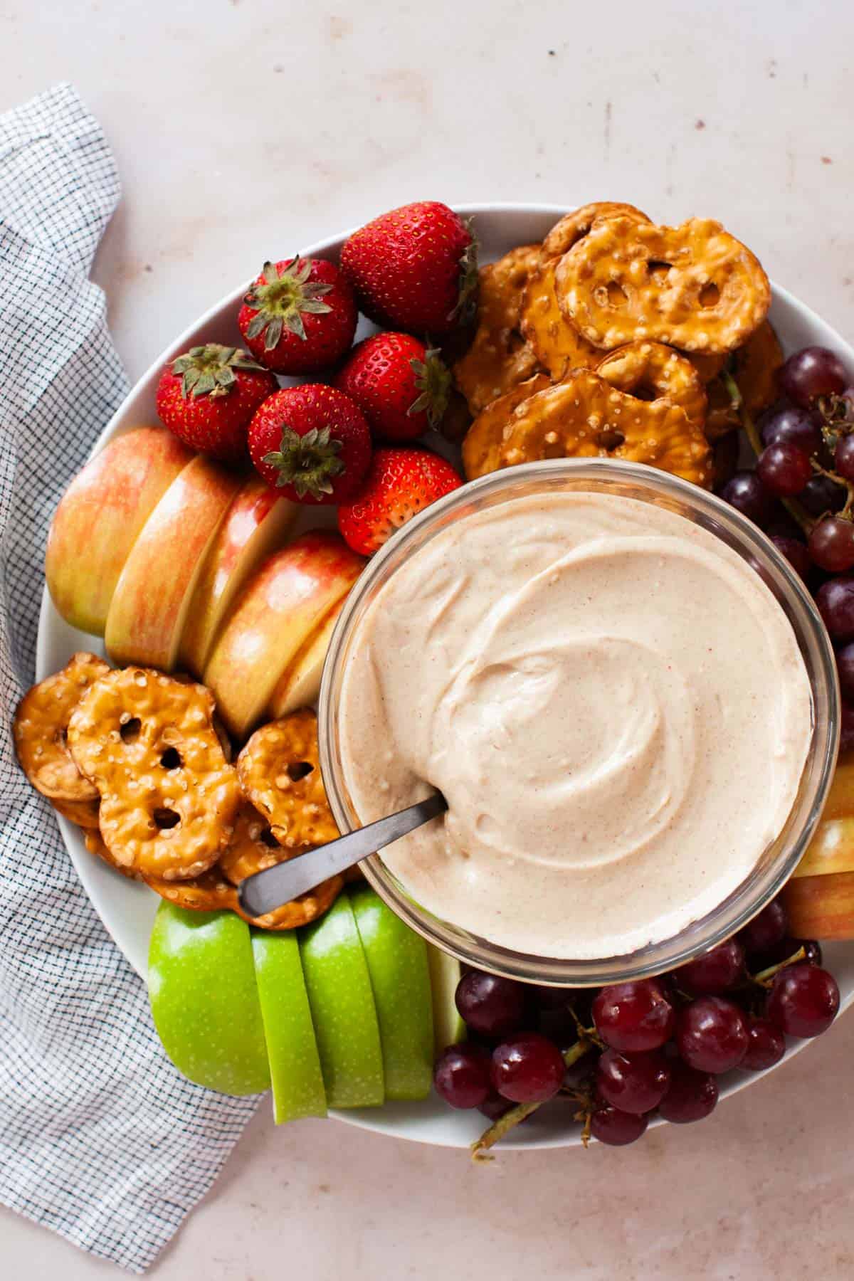 Fruit dip in a glass bowl surrounded by a platter of assorted fruits and pretzels.