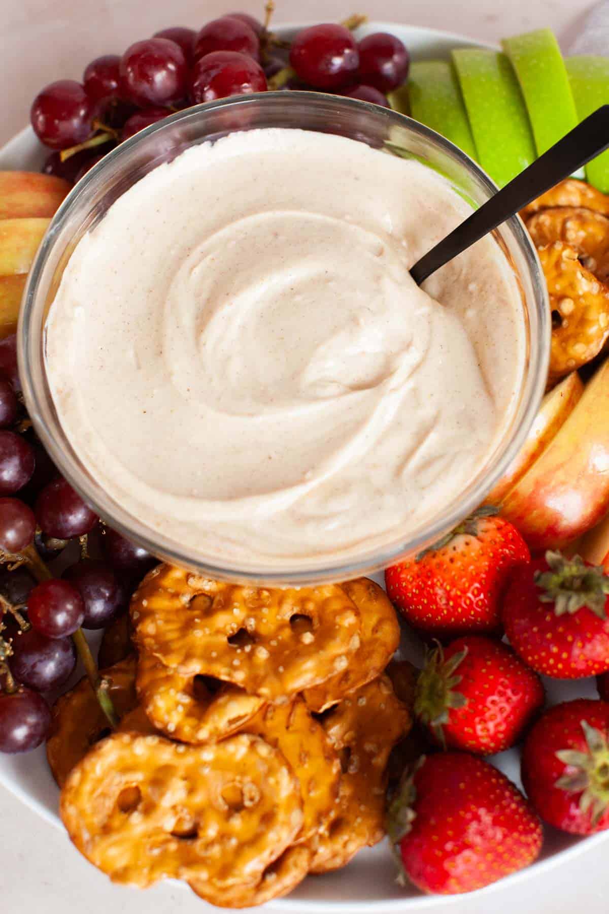 Fruit dip in a glass bowl surrounded by an assortment of grapes, apples and strawberries.