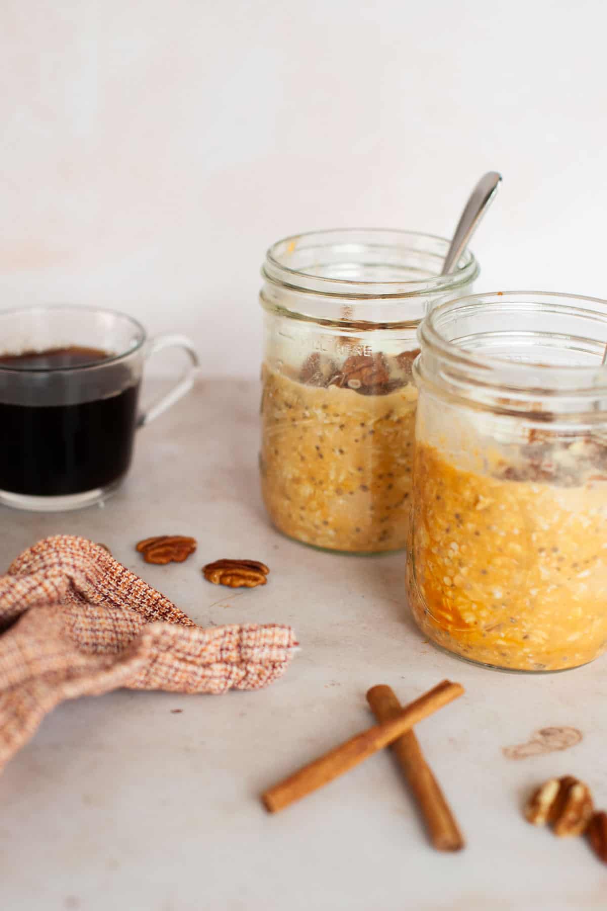 Two mason jars filled with pumpkin overnight oats arranged next to a glass mug with coffee.