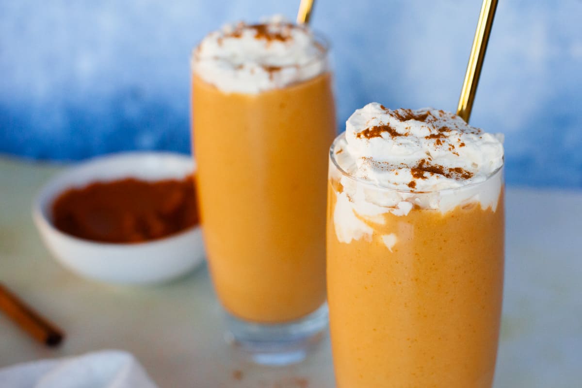 Two pumpkin smoothies topped with cinnamon sprinkled whipped cream and gold straws.