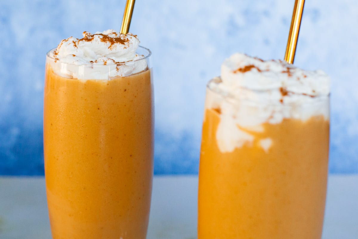 Two pumpkin smoothies topped with cinnamon sprinkled whipped cream and gold straws.