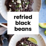 A plate of creamy refried black beans garnished with crumbled cheese, fresh chopped herbs, and a lime wedge, shown from above.