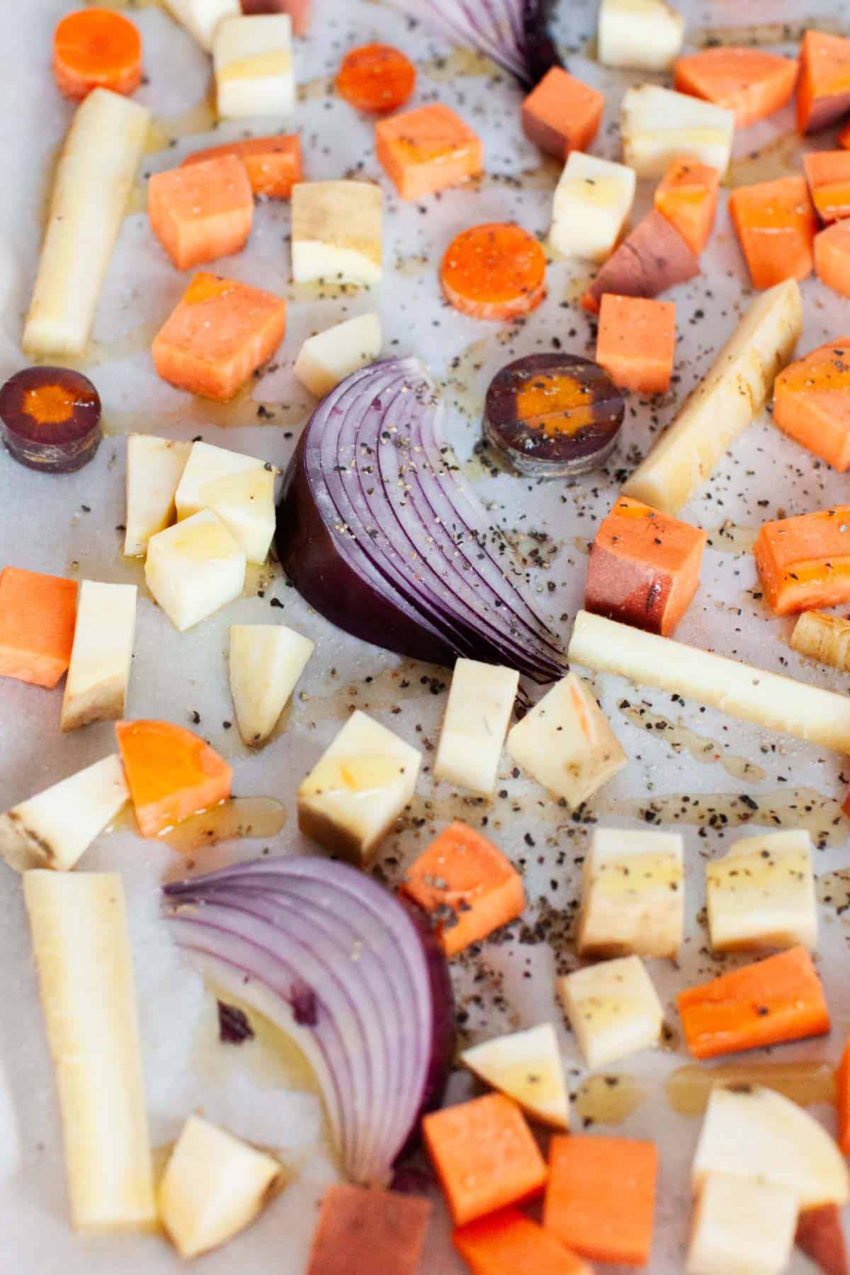A baking sheet with red onion wedges, carrots, yams and white sweet potatoes seasoned with olive oil, salt and pepper.