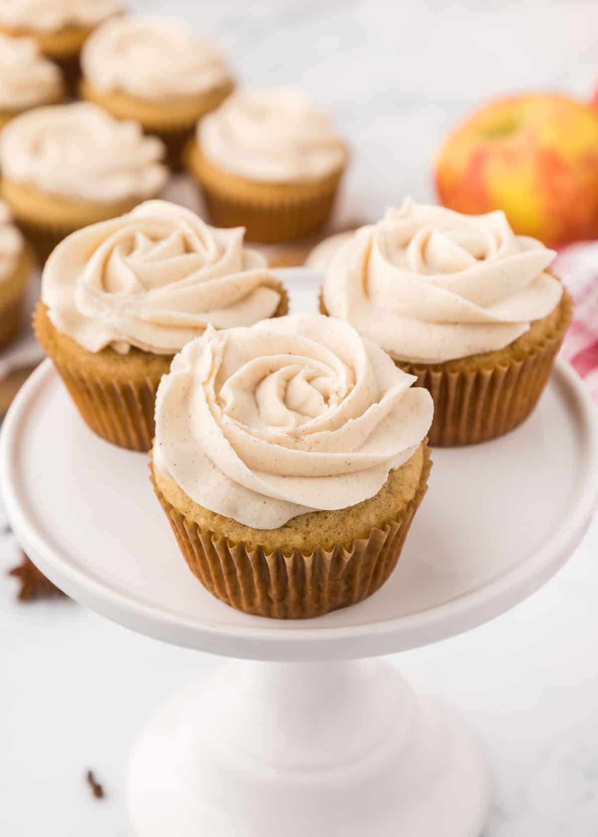 Three apple spice cupcakes with cinnamon frosting on a white cake stand.