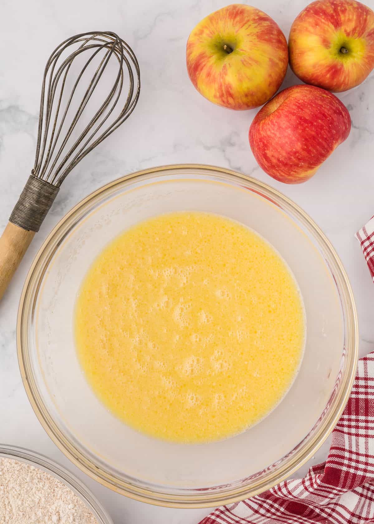 A bowl of whisked eggs on a marble countertop next to apples.