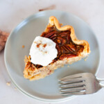 A slice of pecan pie on a plate with whipped cream sprinkled with pecan pieces.