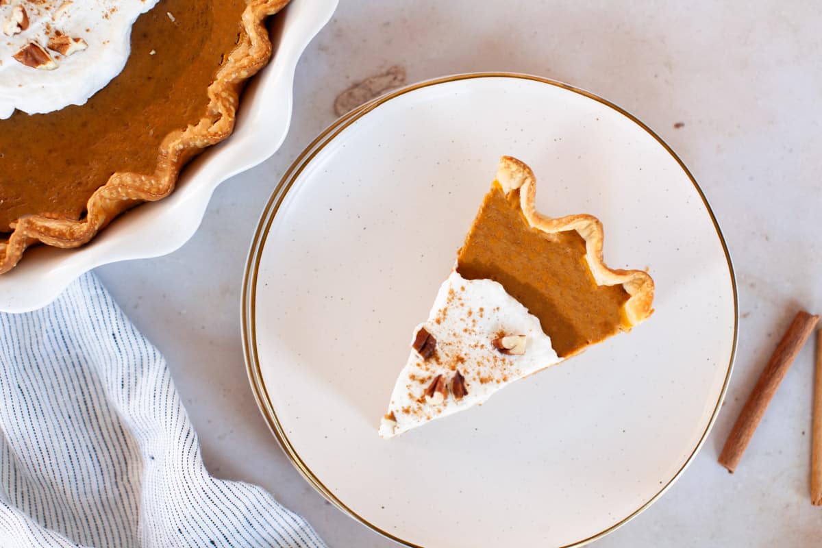 A slice of pumpkin pie on a plate next to two cinnamon sticks.