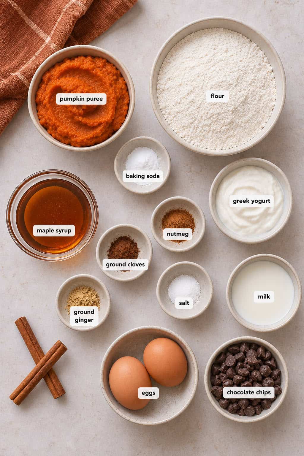 Assorted baking ingredients in bowls, perfect for making pumpkin chocolate chip muffins, including pumpkin puree, flour, greek yogurt, eggs, spices, milk, maple syrup, chocolate chips, baking soda, and salt&mdash;all arranged on a light surface.