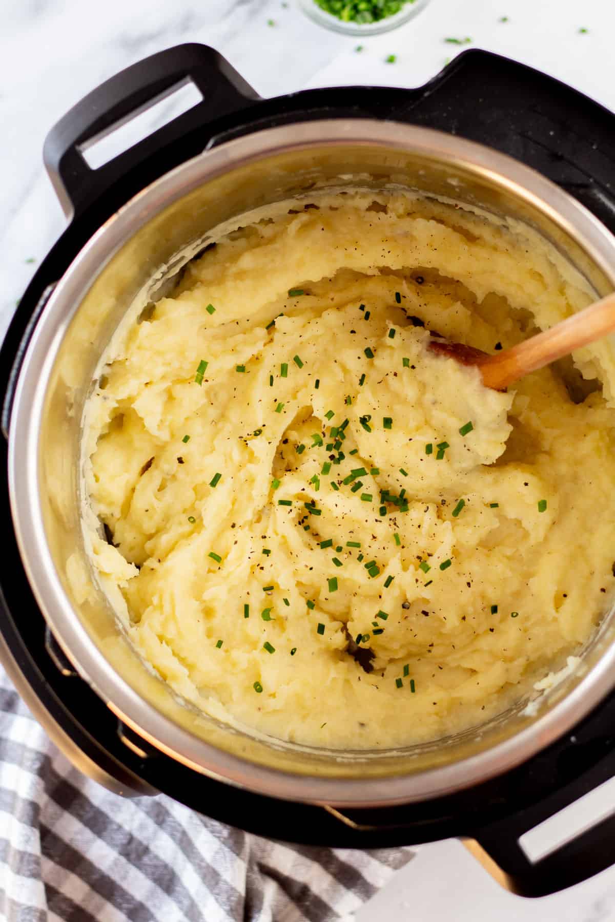 A pot of creamy instant pot mashed potatoes garnished with chopped herbs, with a wooden spoon, on a white background.