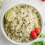 A bowl of cilantro cauliflower lime rice with lime wedges and cherry tomatoes on a marble countertop.