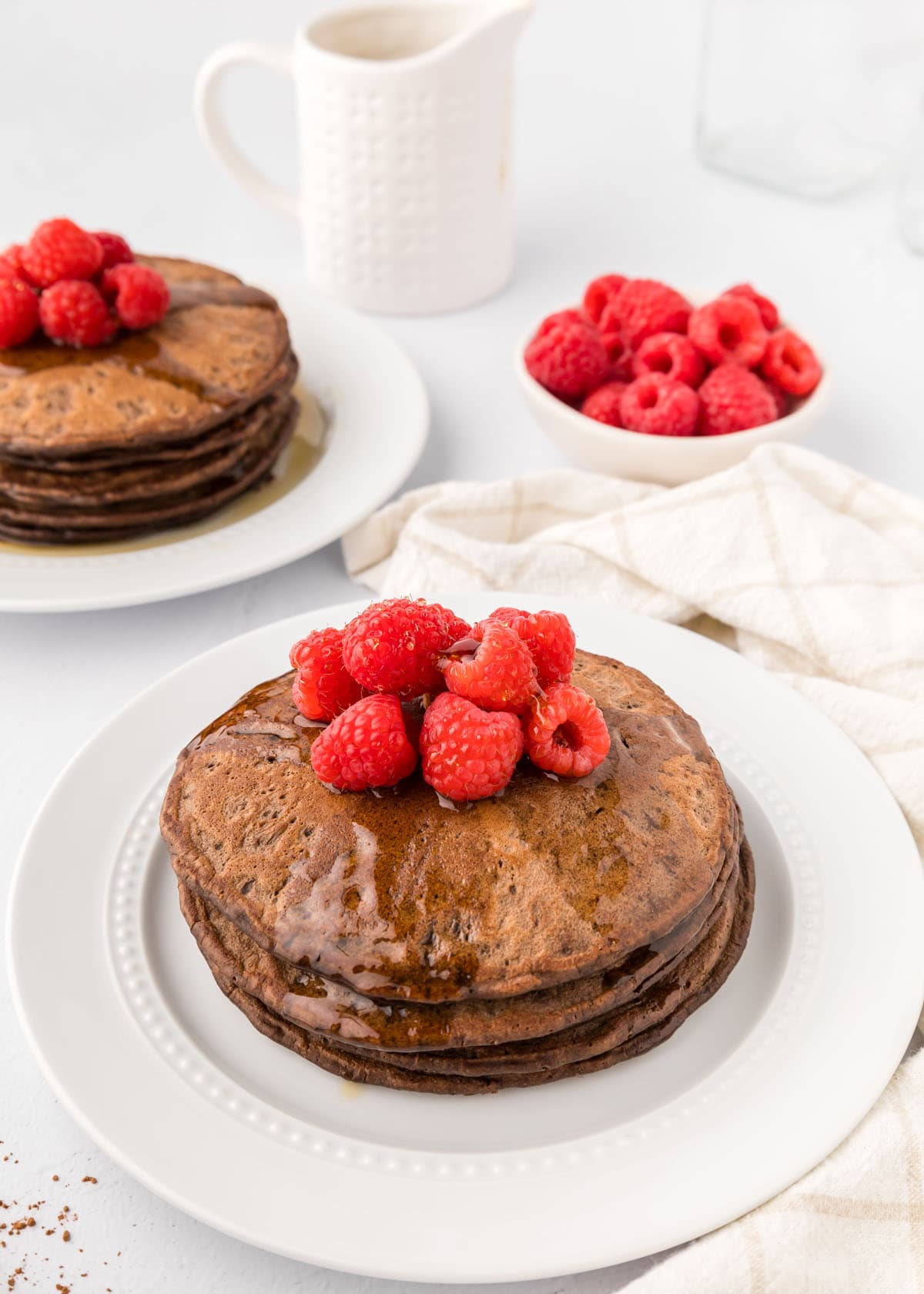 Two plates of chocolate pancakes topped with raspberries and syrup.