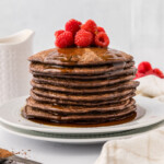 A stack of chocolate pancakes topped with raspberries and syrup on a white plate.