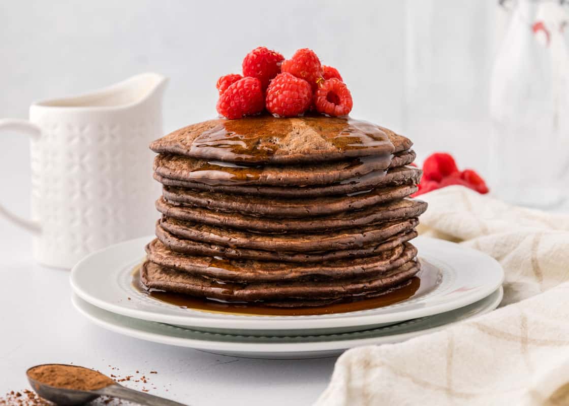 A stack of chocolate pancakes topped with raspberries and syrup on a white plate.