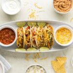 A plate of four chicken fajita tacos with lime wedges. Surrounding the plate are bowls of salsa, shredded cheese, sour cream, beans, guacamole, and tortilla chips. An additional bowl with rice is visible.