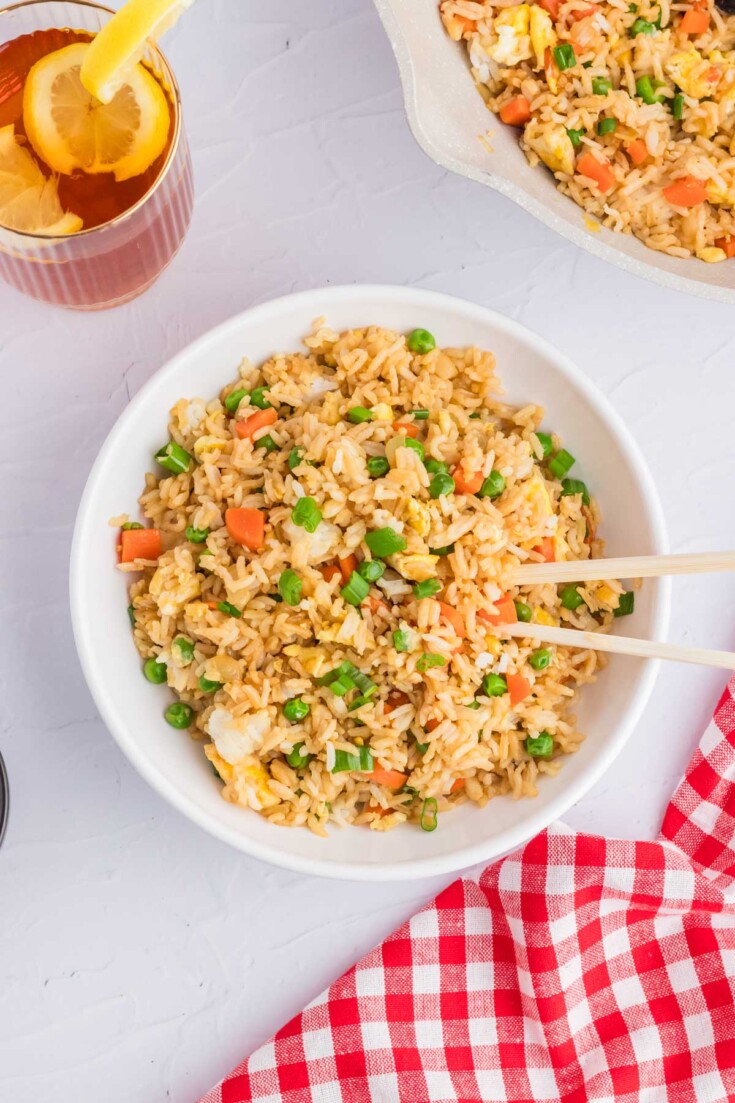A bowl of fried rice with peas, carrots, and egg, accompanied by chopsticks and a lemon iced tea on a checkered red napkin.