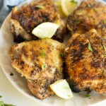 Grilled chicken thighs on a white plate garnished with lemon wedges and parsley, with a blue cloth in the background.