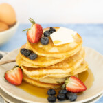 A stack of pancakes topped with butter, blueberries, and a strawberry, drizzled with syrup on a plate, with eggs in the background.