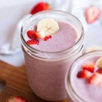 Two jars filled with pink fruit smoothie topped with banana slices and strawberry pieces, placed on a wooden surface with additional strawberries in the background.