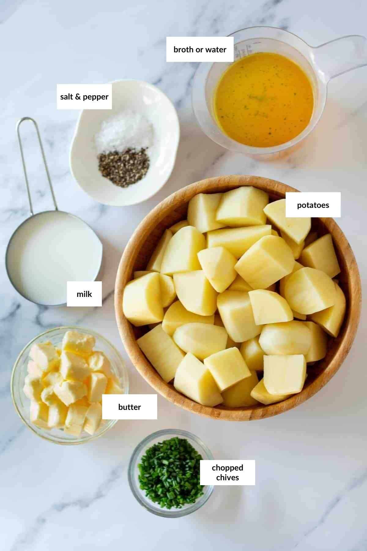 Top-down view of Instant Pot mashed potatoes ingredients: chopped potatoes, butter, milk, salt and pepper, broth or water, and chopped chives in bowls on a white surface, each labeled with text.