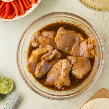 Raw chicken thighs marinating in a glass bowl next to sliced onions, peppers, lime halves, a grater, and tortillas on a table.