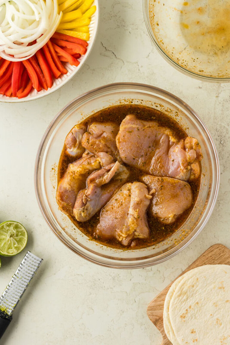 Raw chicken thighs marinating in a glass bowl next to sliced onions, peppers, lime halves, a grater, and tortillas on a table.
