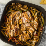 A dish of grilled chicken slices mixed with bell peppers and onions in a black square pan, surrounded by bowls of guacamole, salsa, shredded cheese, and tortilla on a light-colored table.