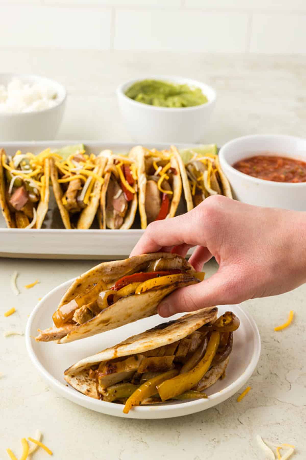 A person holds a taco with grilled vegetables and cheese over a plate, with more tacos, salsa, guacamole, and rice in the background.