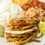 A plate with three fajitas filled with sliced bell peppers and onions, served with a side of white rice, refried beans, tortilla chips, salsa, and avocado slices.