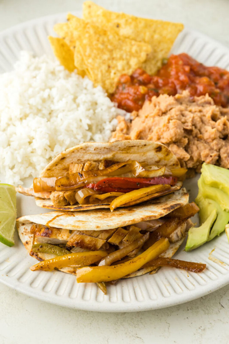 A plate with three fajitas filled with sliced bell peppers and onions, served with a side of white rice, refried beans, tortilla chips, salsa, and avocado slices.