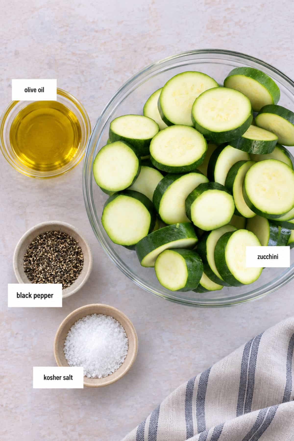 A glass bowl of sliced zucchini, perfect for a saut&eacute;ed zucchini recipe, sits alongside small bowls of olive oil, black pepper, and kosher salt on a light surface next to a striped cloth.