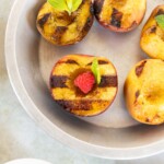 Grilled peach halves garnished with mint leaves and a raspberry, served in a round metal dish. A small bowl of vanilla ice cream is visible nearby.