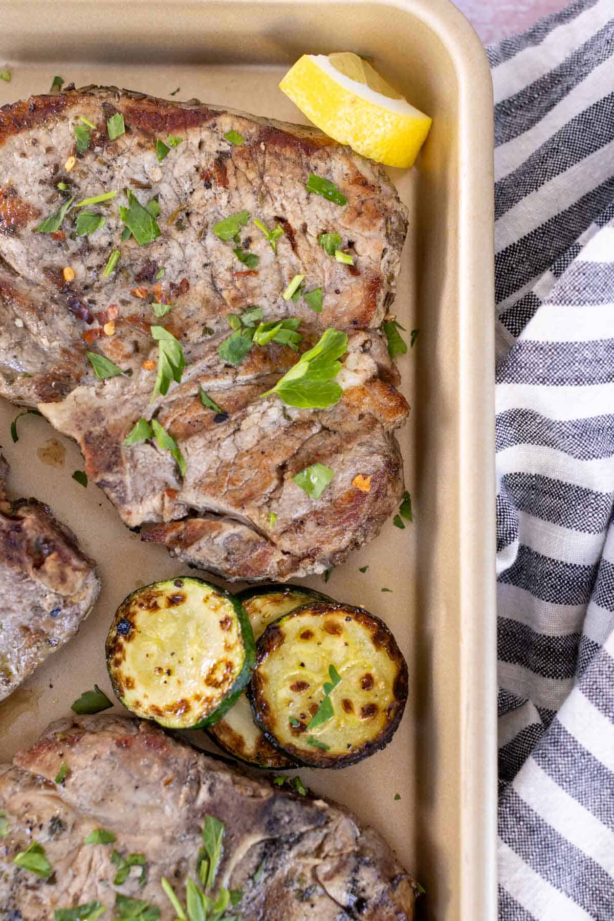 Grilled pork chops with fresh herbs and served with grilled zucchini slices and a lemon wedge on a metal tray. A striped cloth is partially visible on the side.