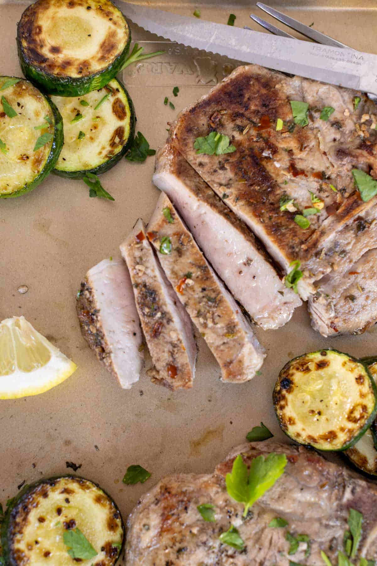 Slices of grilled pork chop and grilled zucchini rounds are on a plate. A lemon wedge and a knife are nearby. Parsley is sprinkled as garnish.