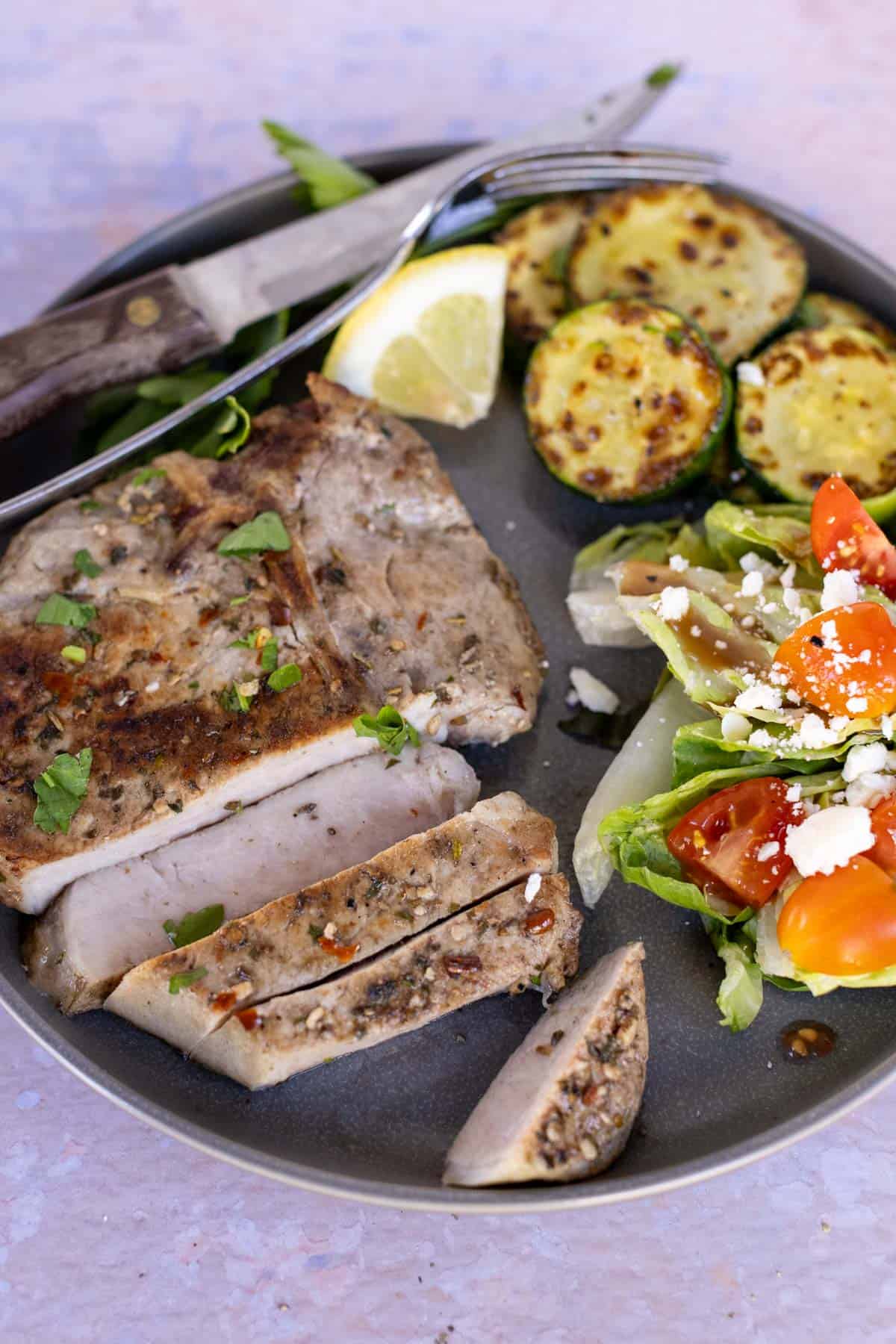 A plate with sliced seasoned pork chop, saut&eacute;ed zucchini, and a side salad with tomatoes, onions, and feta cheese. A knife and fork are on the plate's edge; a lemon wedge is also present.